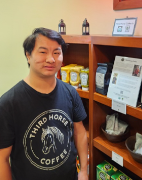 Josiah standing beside a display of Third Horse Coffee at the White Horse Village market.